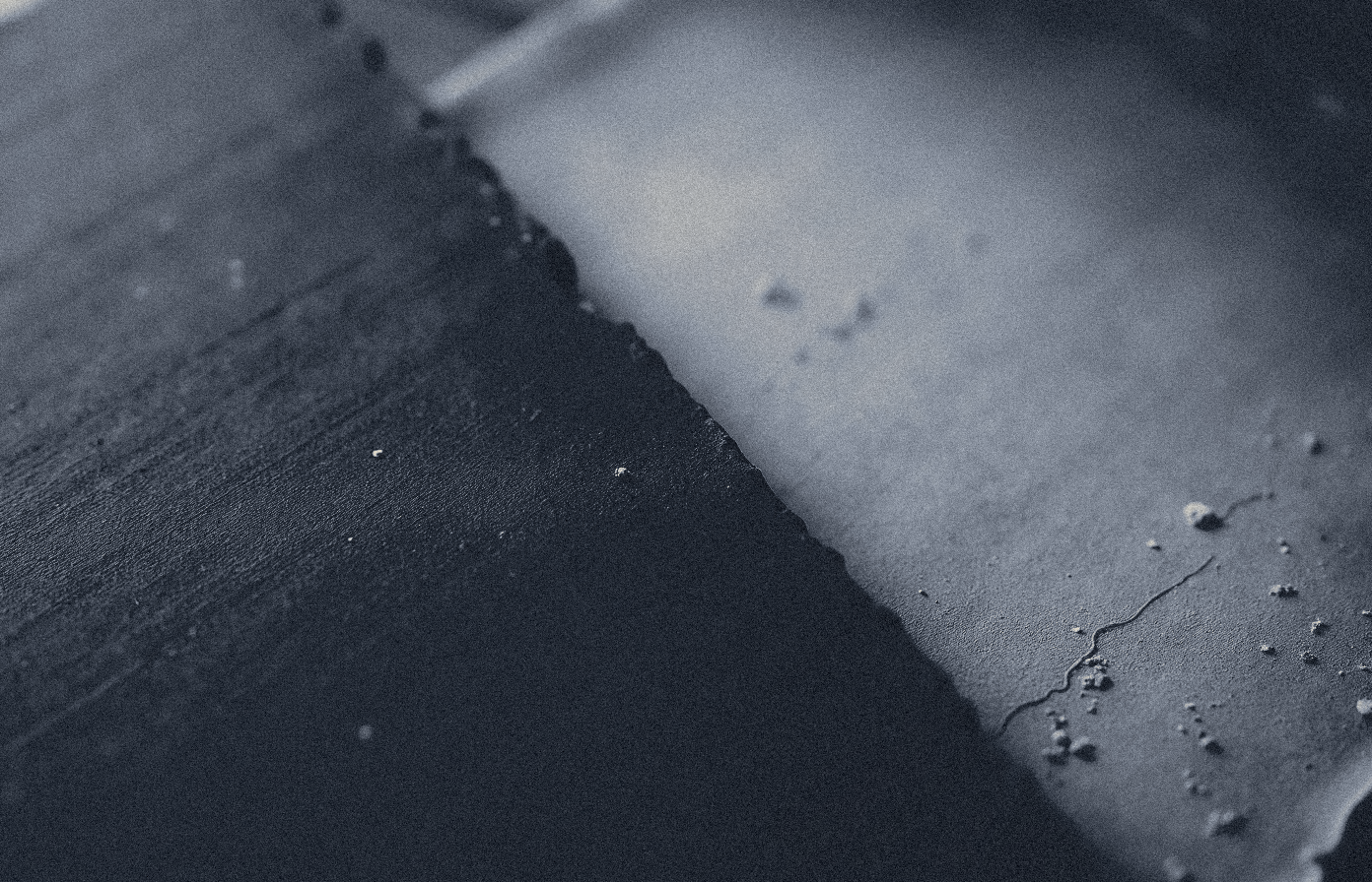 Macro image of the edge of a dark wax writing tablet beside the edge of a blank pale sheet, both empty, evoking two eras of Greek writing separated by centuries.