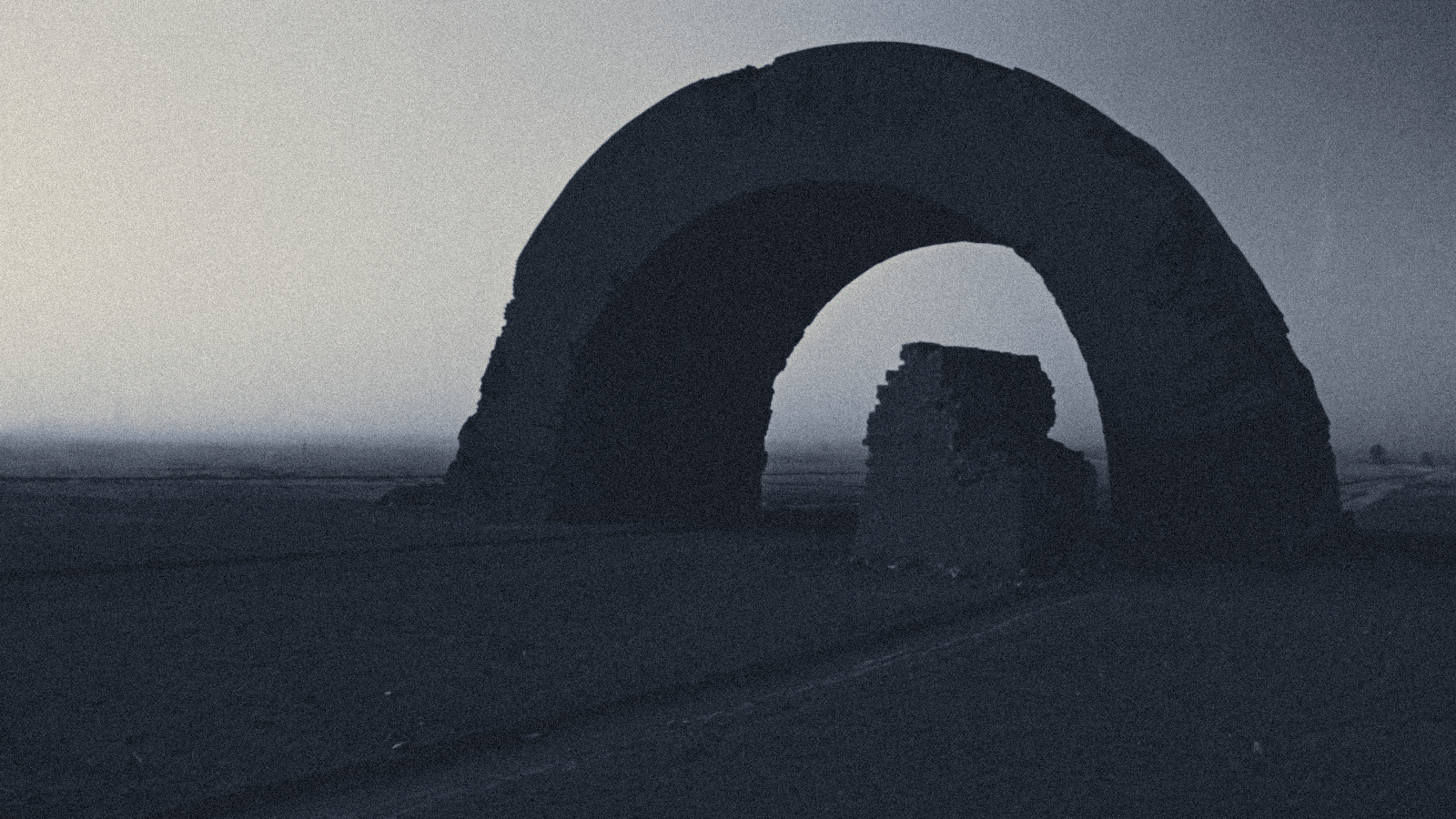 Silhouette of a single vast ruined brick arch standing alone on a flat hazy plain at dusk, a broken stub of wall beneath it.